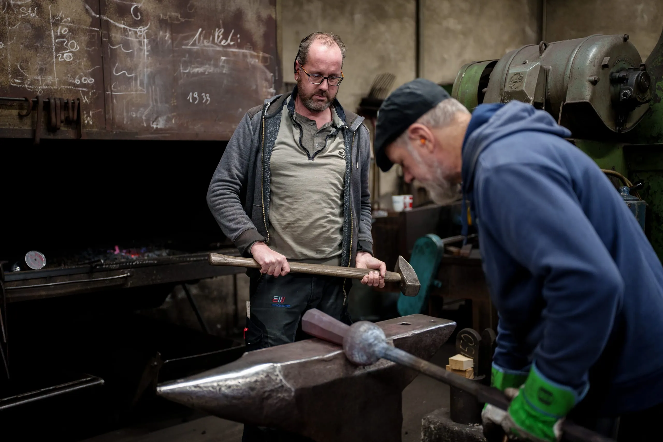 Zwei Hammerschmiede arbeiten synchron an einem Glockenklöppel, um die Form für die Glocken der Notre-Dame zu präzisieren. Mit kräftigen Schlägen und meisterhafter Handwerkskunst formen sie das Metall, das später den ehrwürdigen Klang der historischen Glocken erzeugen wird.